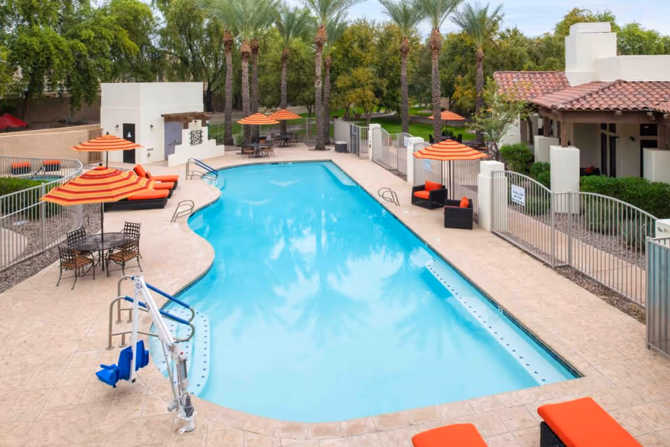 A rectangular outdoor swimming pool surrounded by orange-cushioned lounge chairs, striped umbrellas, tables, and palm trees.