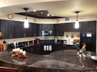 A spacious kitchen with dark wood cabinets, black appliances including a refrigerator, microwave, and stove. The kitchen features a large curved granite countertop with decorative candles and a rooster figurine. Two pendant lights hang from the ceiling, and the walls have decorative plates and words displayed near the ceiling.