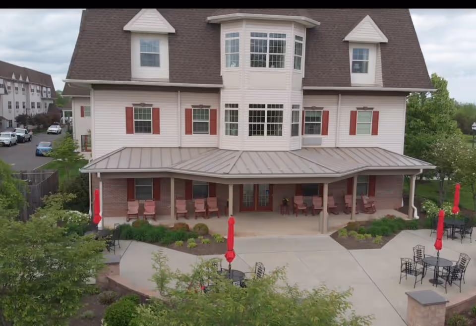 Exterior view of a multi-story assisted living facility building with a covered porch area featuring several chairs. In front of the building, there is a paved patio area with round tables, chairs, and red umbrellas. Surrounding the patio are landscaped garden beds and greenery.