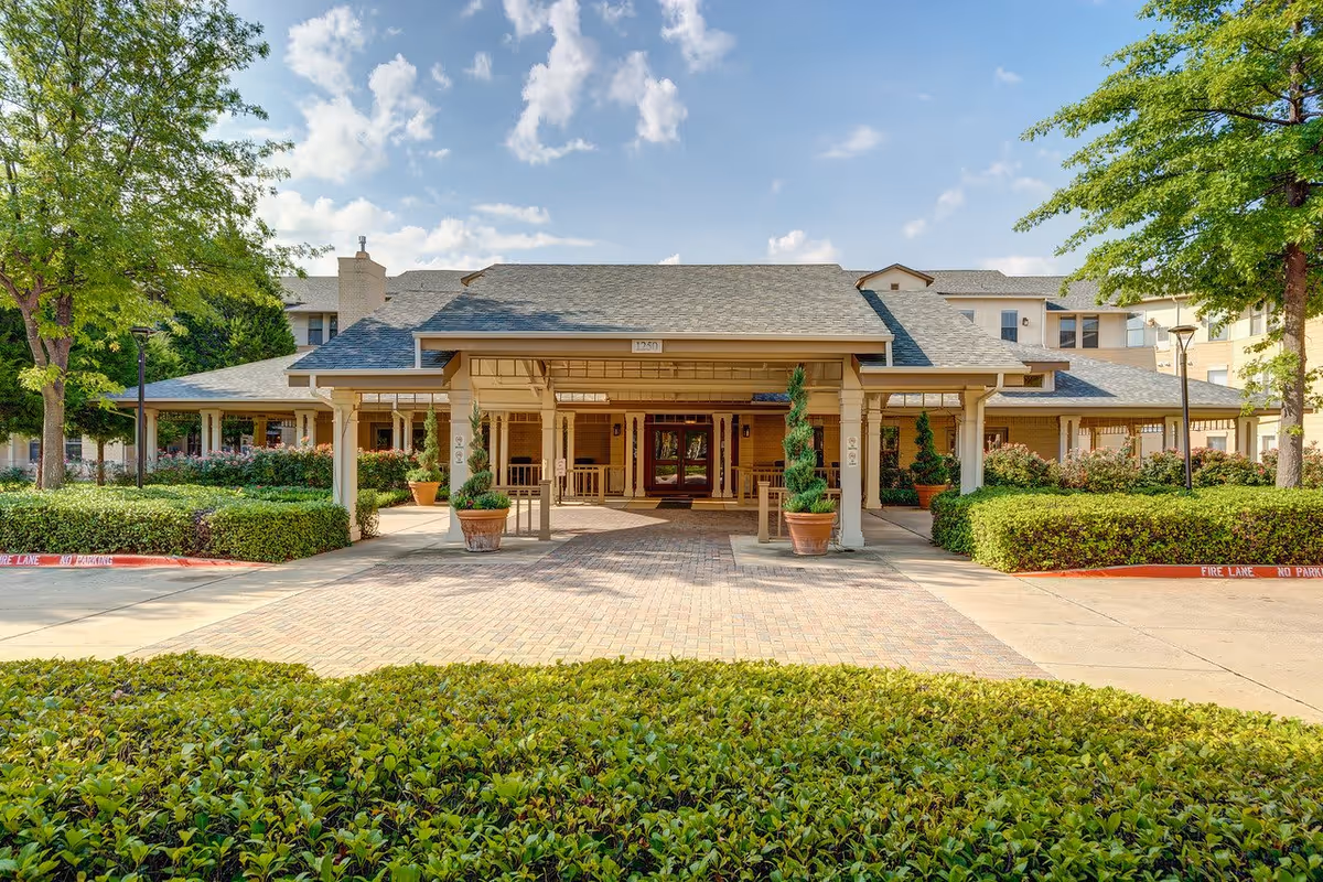Front exterior view of Town Village Crossing facility showing a covered entrance with potted plants, a paved driveway, green bushes, and trees under a partly cloudy sky.