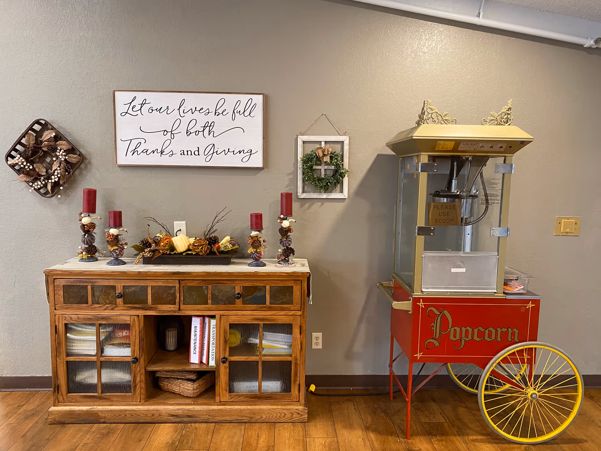 A cozy interior corner featuring a wooden cabinet decorated with autumn-themed items including red candles, pinecones, and a small pumpkin centerpiece. Above the cabinet is a wall sign that reads 'Let our lives be full of both Thanks and Giving' and a small framed wreath. To the right of the cabinet is a vintage-style popcorn machine with red and yellow accents on wooden flooring.