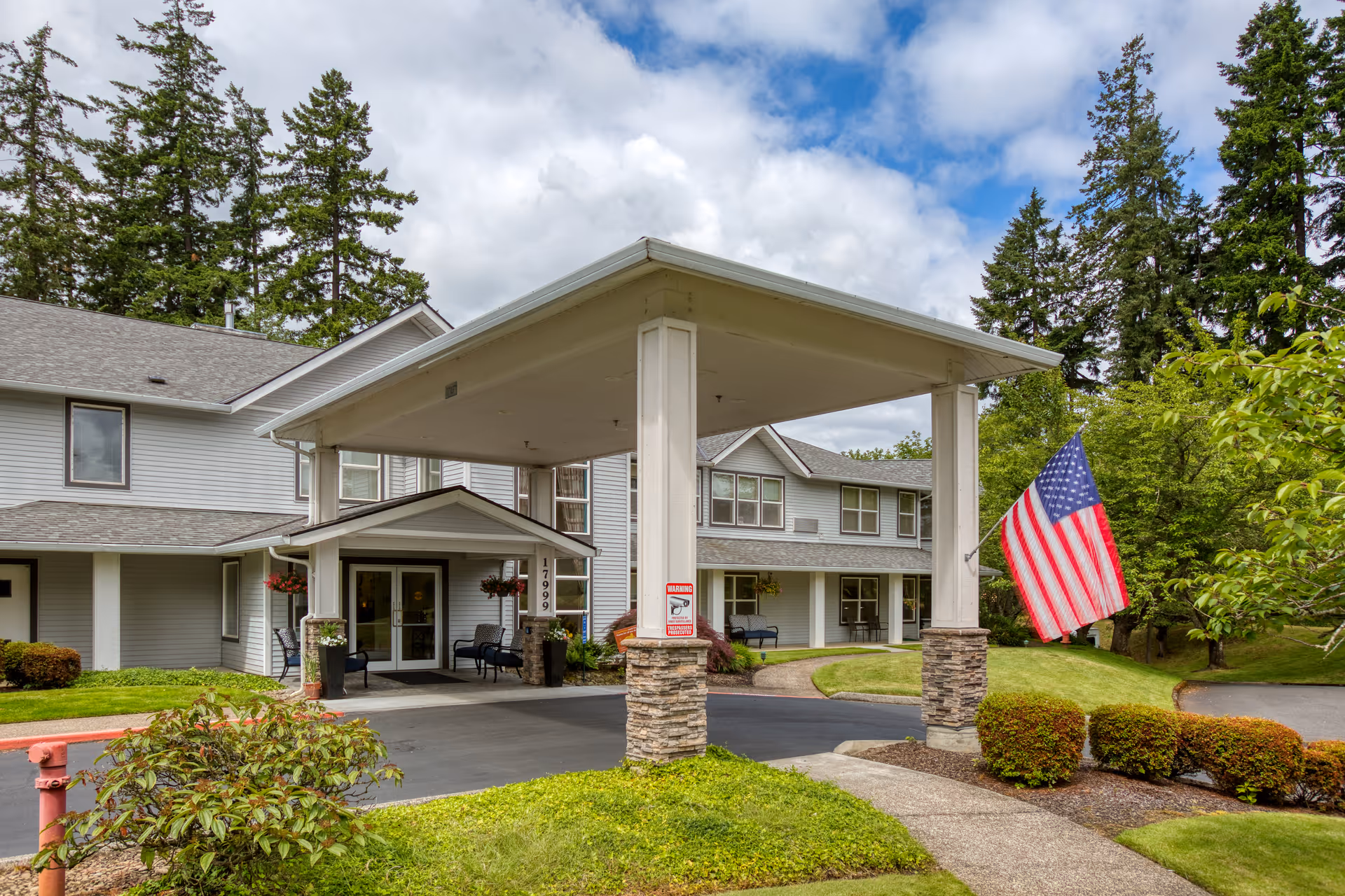 Exterior view of Homewood Assisted Living facility showing the entrance with a covered driveway, an American flag on the right, well-maintained landscaping, and a two-story building with gray siding and multiple windows under a partly cloudy sky.