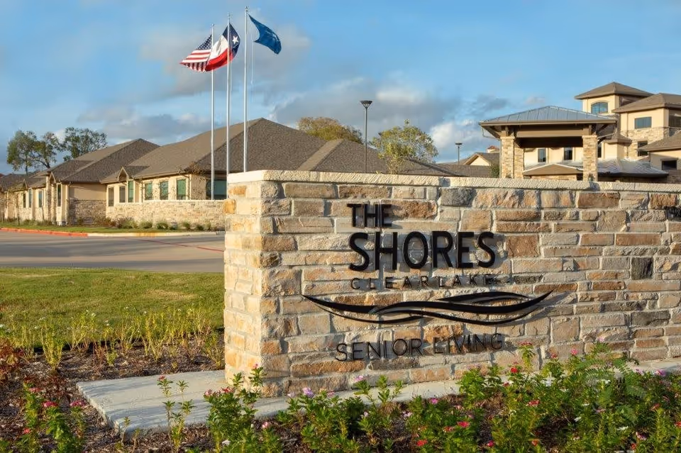 Stone sign at the entrance of The Shores at Clear Lake senior living facility with three flagpoles flying the American, Texas, and a blue flag, with the facility buildings and landscaping in the background under a partly cloudy sky.