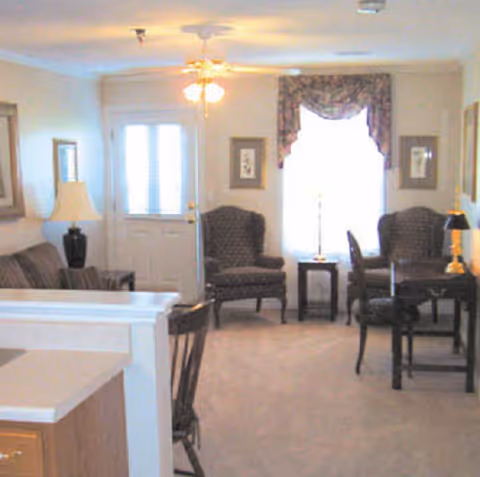 A cozy living room area with two patterned armchairs, a small table with a lamp between them, a sofa, a ceiling fan with lights, and a door with windows letting in natural light. The room has framed artwork on the walls and a window with floral valance curtains.