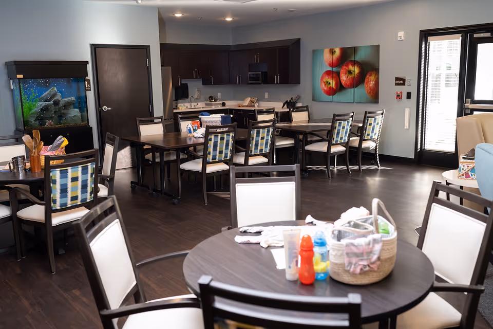 Interior view of a communal dining and activity area in a senior living facility. The room features multiple tables and chairs with patterned cushions, a large fish tank on the left, a kitchenette with dark cabinets in the background, and a colorful painting of apples on the wall. Various items including bottles and a basket are placed on the tables. There is a door and large windows letting in natural light on the right side.