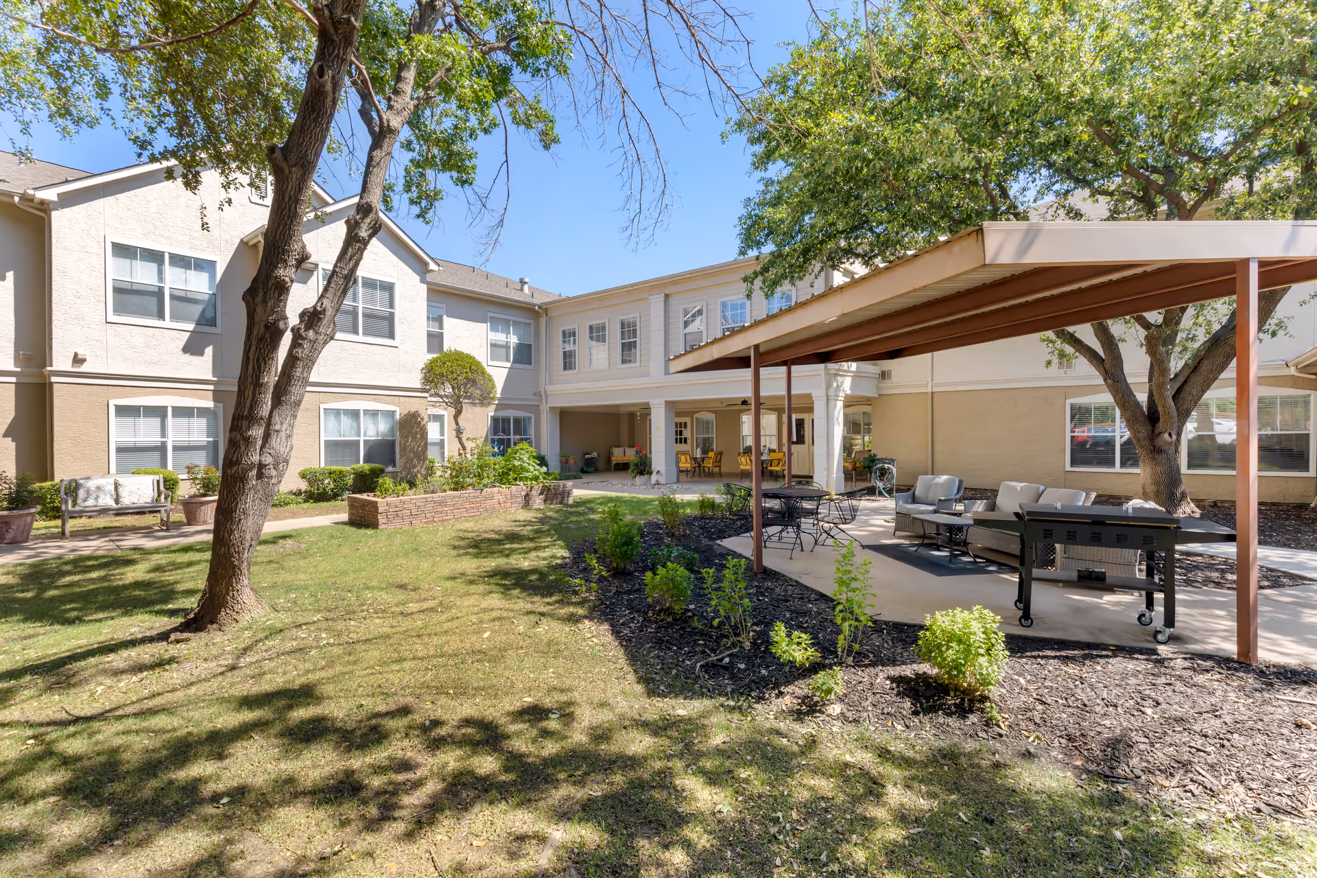 Outdoor courtyard area of a senior living facility with green grass, trees, and shrubs. There is a covered patio with outdoor seating including cushioned chairs and a table, as well as a grill. The building surrounding the courtyard is two stories with multiple windows and beige exterior walls.