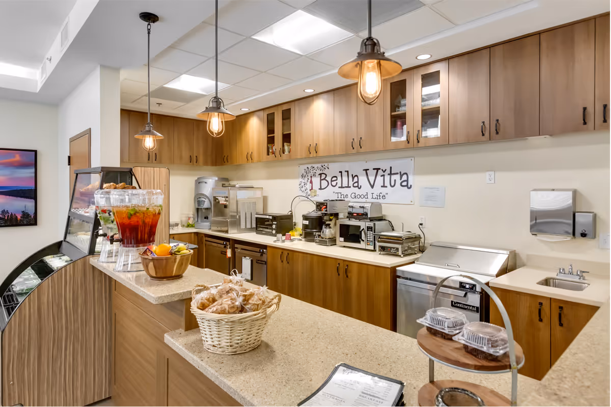 Interior view of a serving counter and kitchenette with pendant lights, beverages, pastries, and a 'Bella Vita' sign.