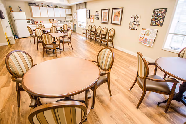 A bright dining area with round wooden tables and striped cushioned chairs. The room has a wooden floor, a row of chairs lined up against the wall, framed artwork, and a kitchenette with white cabinets, a refrigerator, and a microwave in the background.