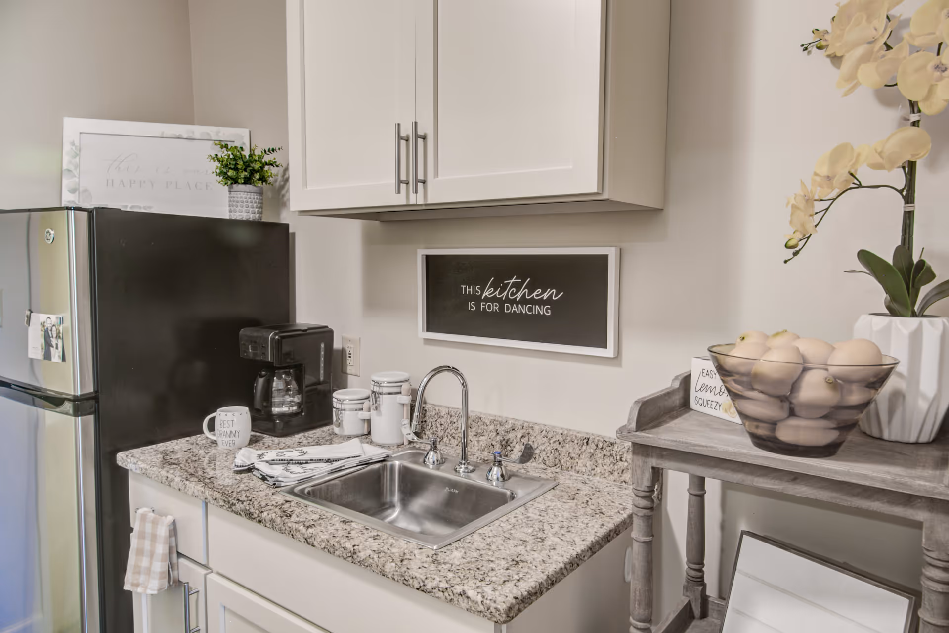 A small kitchen area featuring a granite countertop with a stainless steel sink and faucet. Above the sink is a white cabinet and a framed sign that reads 'This kitchen is for dancing.' To the left is a black refrigerator with a coffee maker and mugs on the counter. To the right is a wooden side table with a glass bowl filled with apples and a white orchid plant in a vase.