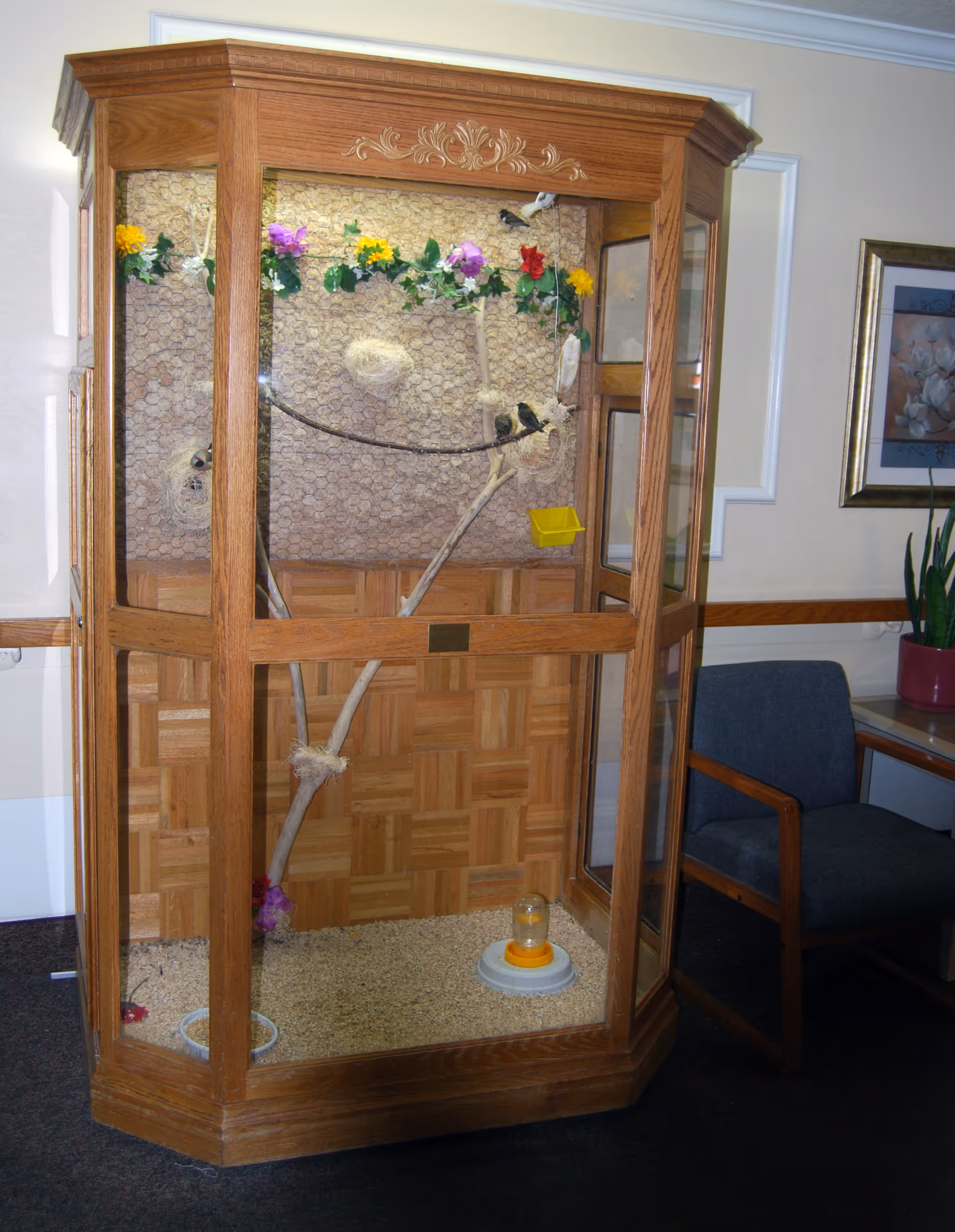 A wooden and glass birdcage with decorative flowers and branches inside, placed in a room with a blue upholstered chair and a framed floral painting on the wall.