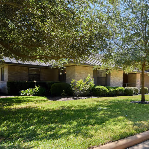 Exterior view of a single-story brick building with a sloped roof, surrounded by green grass, bushes, and trees providing shade on a sunny day.