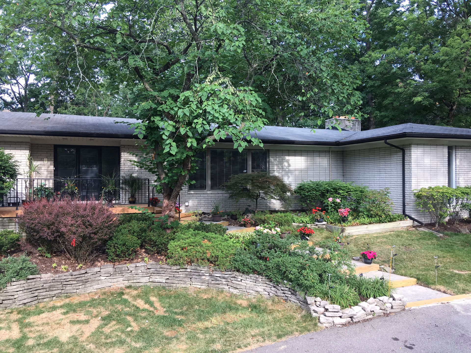 Front exterior view of a single-story brick building with a sloped roof, surrounded by a well-maintained garden with various shrubs, flowers, and a large tree in the center. A stone retaining wall borders the garden, and there is a small set of steps leading up to the entrance.