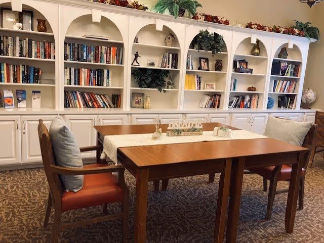 A cozy interior room with a wooden table and four chairs, each with a cushion. Behind the table is a large white built-in bookshelf filled with books, decorative items, and plants. The room has a warm and inviting atmosphere with a carpeted floor and soft lighting.