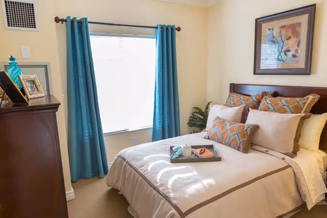 A cozy bedroom with a neatly made bed featuring beige and orange patterned pillows. A tray with a cup and a book rests on the bed. There is a window with teal curtains letting in natural light, a wooden dresser with framed photos, and a framed floral artwork on the wall above the bed.