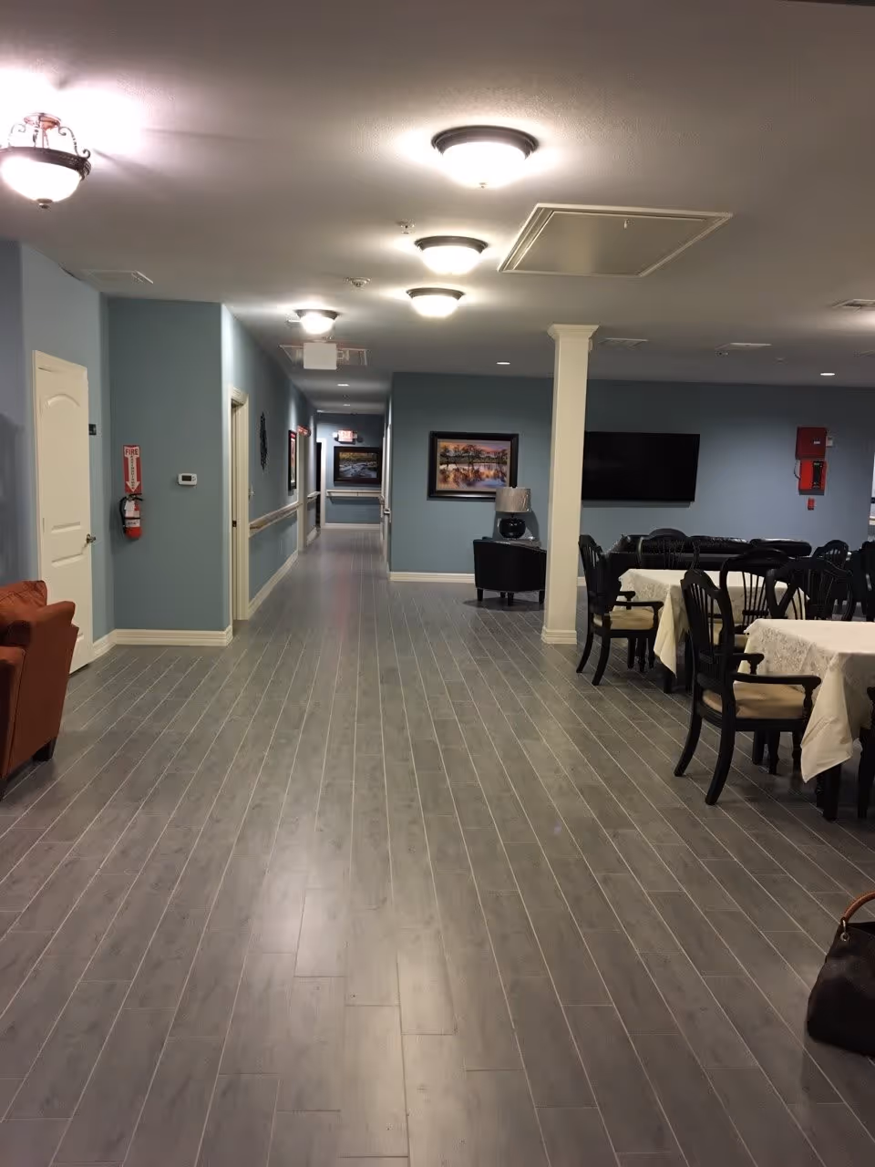 Interior view of a senior living facility hallway with light blue walls and gray wood flooring. On the right side, there is a dining area with tables covered in white tablecloths and black chairs. Further back, there is a black armchair, a lamp, and a wall-mounted TV. The hallway extends into the distance with several doors and framed pictures on the walls. Ceiling lights illuminate the space.
