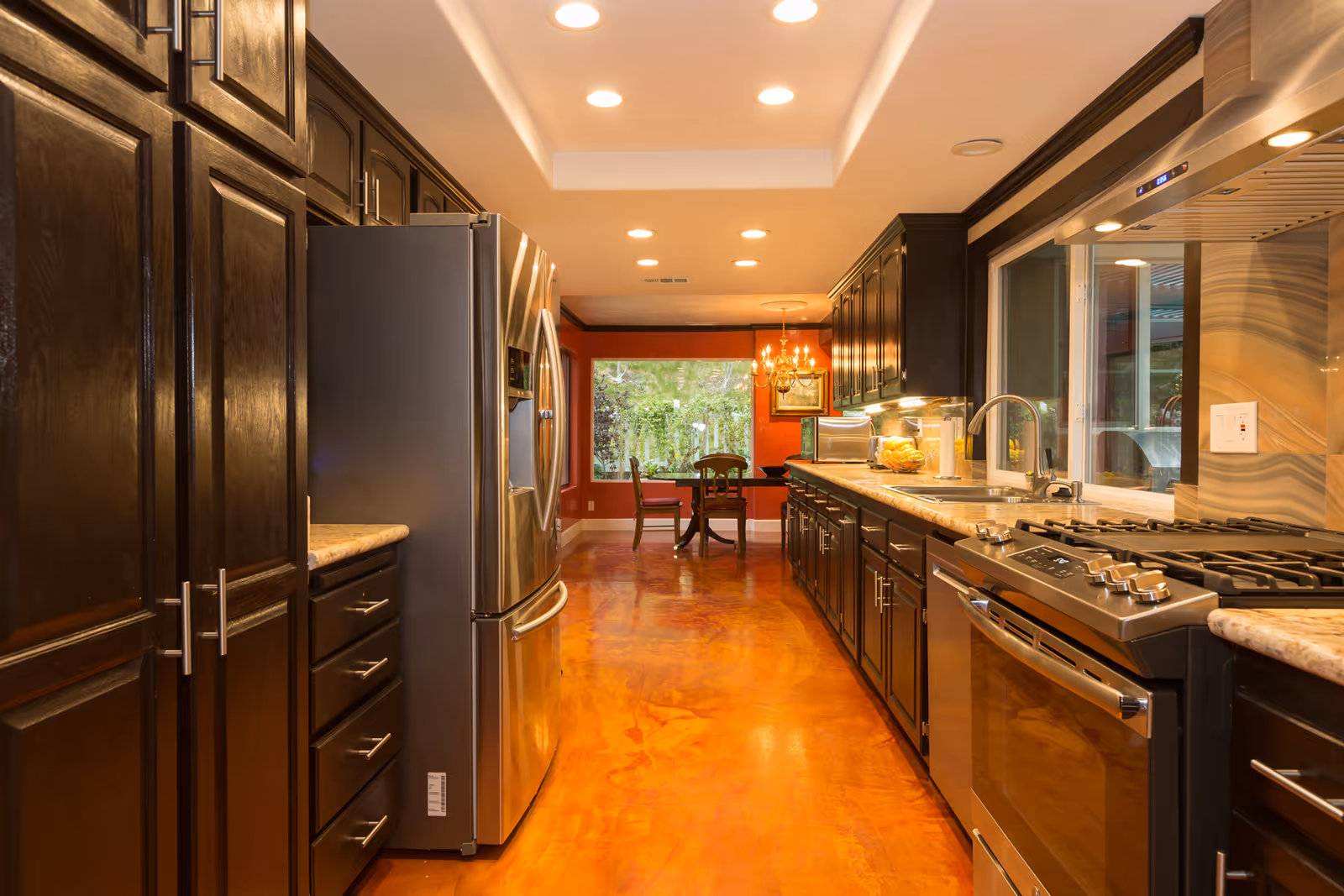 A modern kitchen with dark wooden cabinets, stainless steel refrigerator, stove, and dishwasher. The kitchen has a long countertop with a sink and a window above it. At the far end, there is a dining area with a round table and chairs, a chandelier, and a large window showing greenery outside.