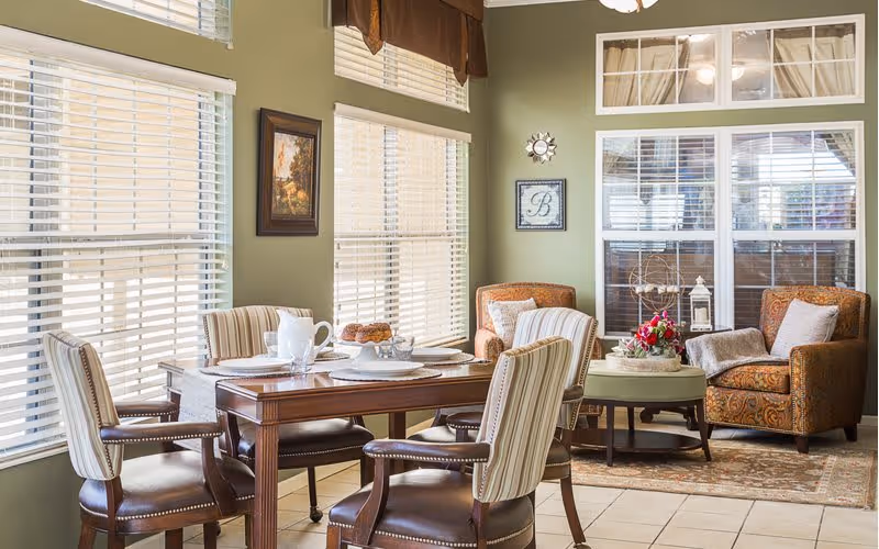 A cozy interior room with a wooden dining table set for tea, surrounded by four striped upholstered chairs. The room has large windows with white blinds and green walls. In the corner, there are two patterned armchairs with cushions and a small round table with a floral arrangement and decorative items. The floor is tiled and there is a rug under the armchairs.