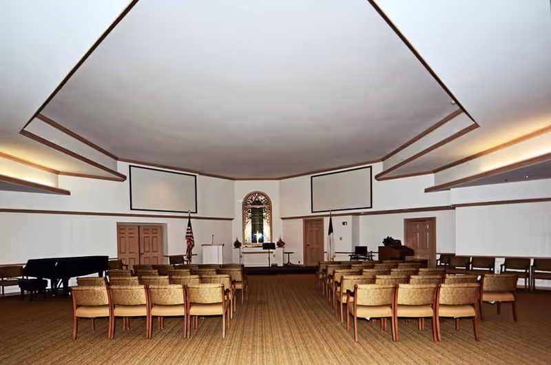 Large chapel-style meeting room with rows of chairs facing a stage with podiums, flags, a piano, and projection screens.