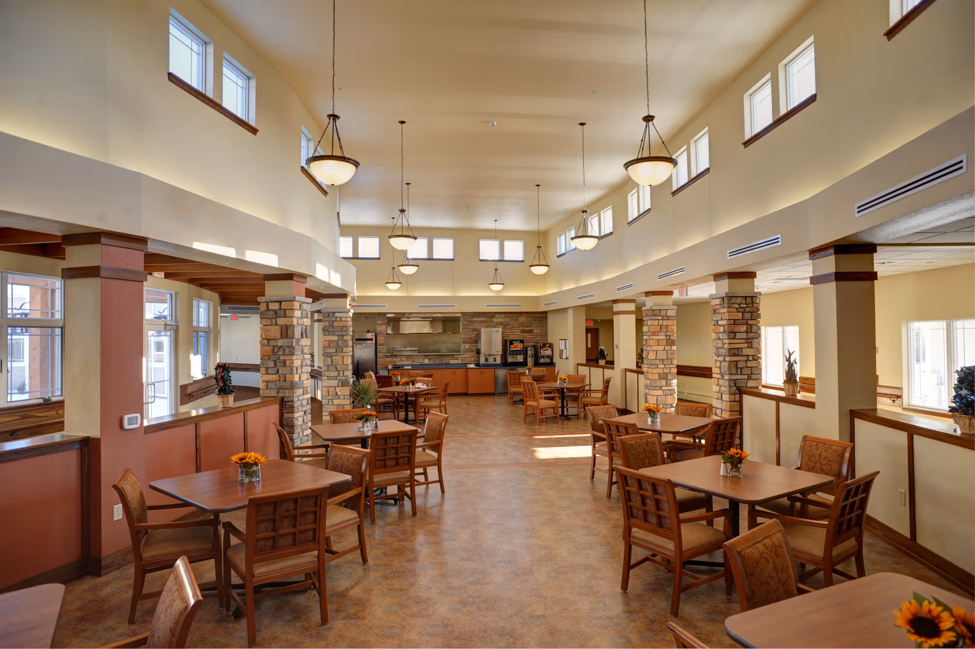 A spacious dining room with multiple wooden tables and chairs arranged neatly. The room features high ceilings with hanging light fixtures and several windows near the ceiling allowing natural light to enter. Stone pillars and warm-toned walls add to the cozy atmosphere. In the background, there is a kitchen area with appliances and a counter.