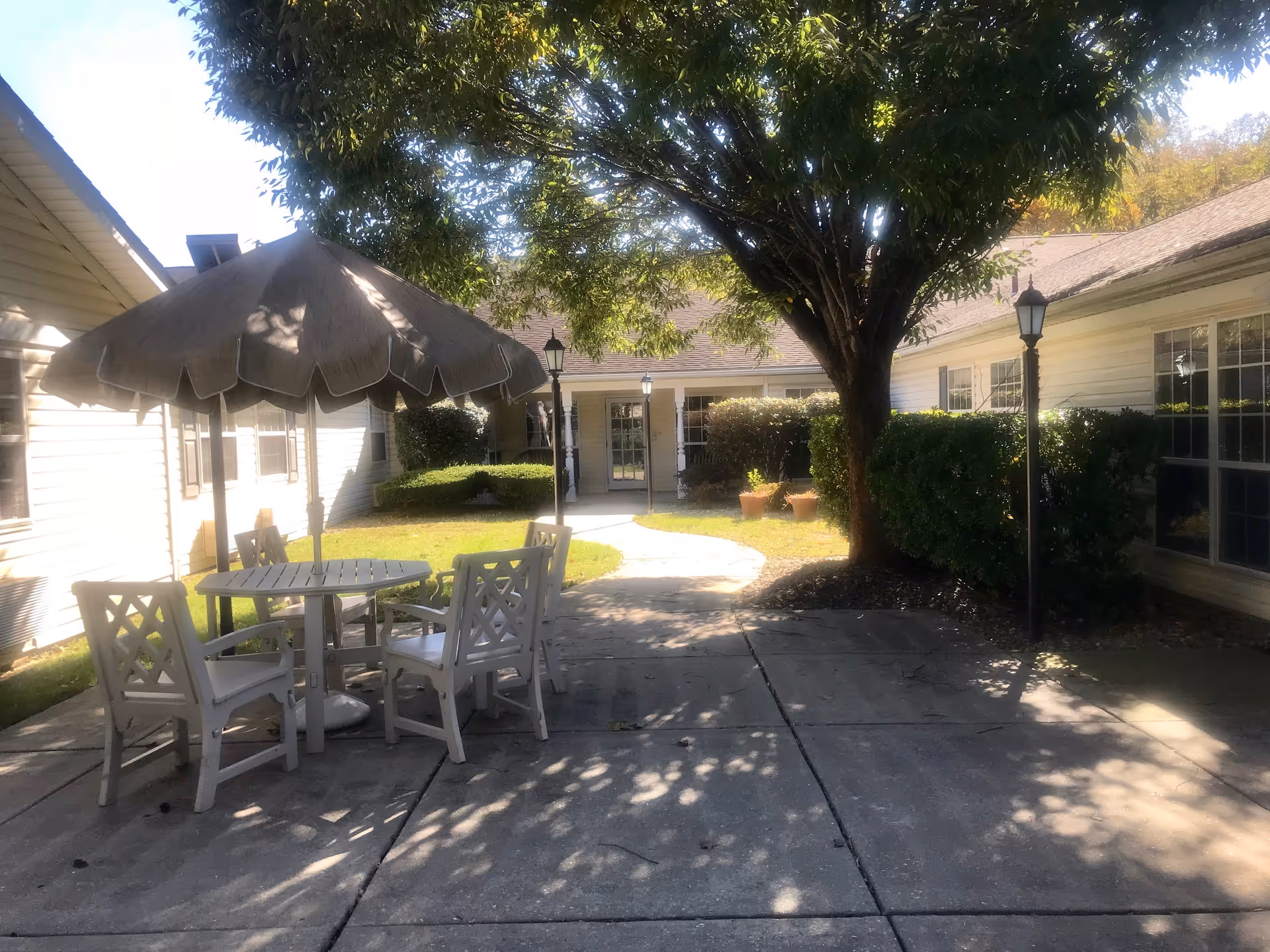 Sunlit courtyard patio with a round table, umbrella and chairs under a tree between single-story buildings.