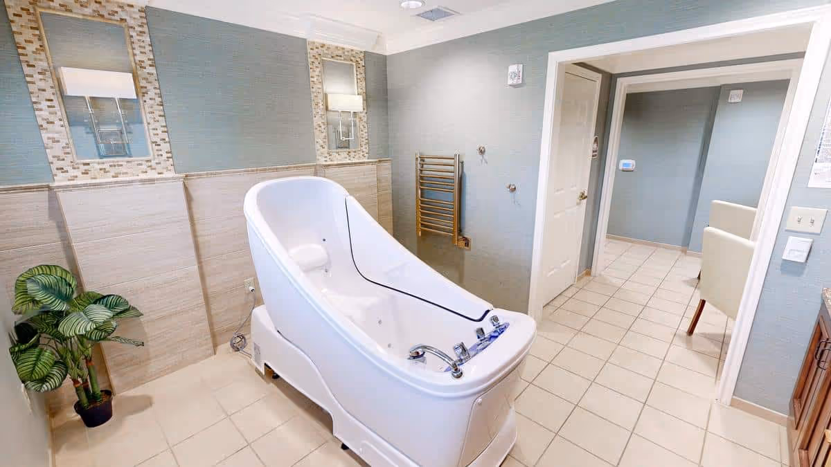 A modern bathroom featuring a large white therapeutic bathtub with built-in controls, light blue and beige tiled walls, two rectangular mirrors with mosaic tile borders, a towel warmer mounted on the wall, and a potted plant in the corner. The room has a tiled floor and an open doorway leading to another area with light-colored furniture.