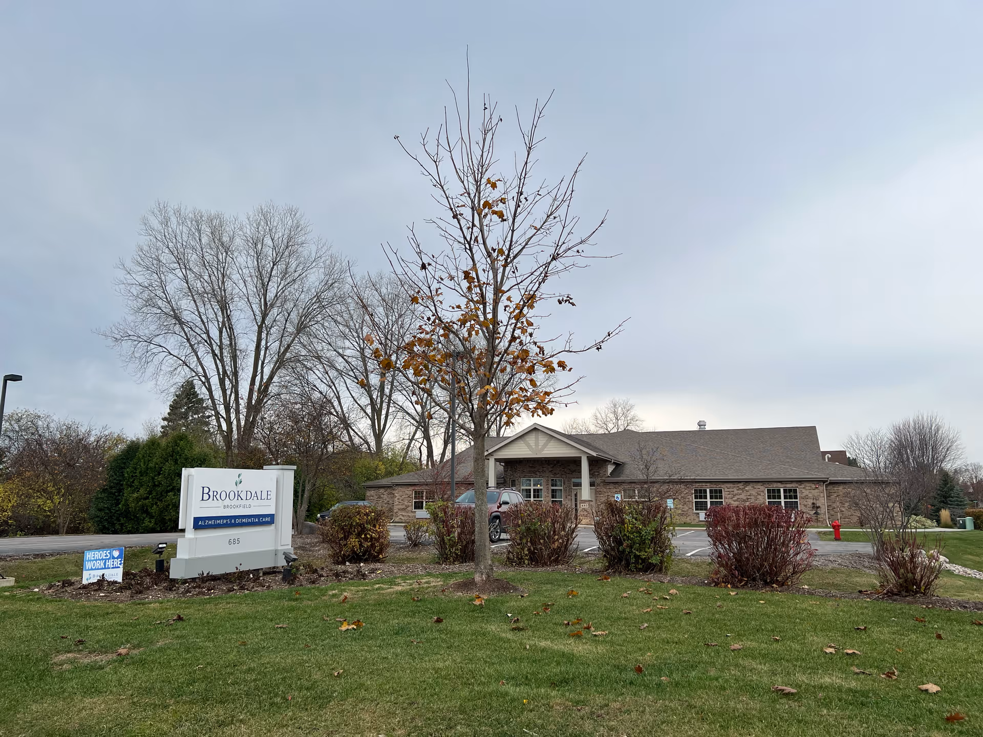 Exterior view of Brookdale Brookfield facility with a brick building, parking lot, and a sign that reads 'Brookdale Brookfield Alzheimer's & Dementia Care'. There is a small tree with some remaining autumn leaves in the foreground and a cloudy sky overhead.