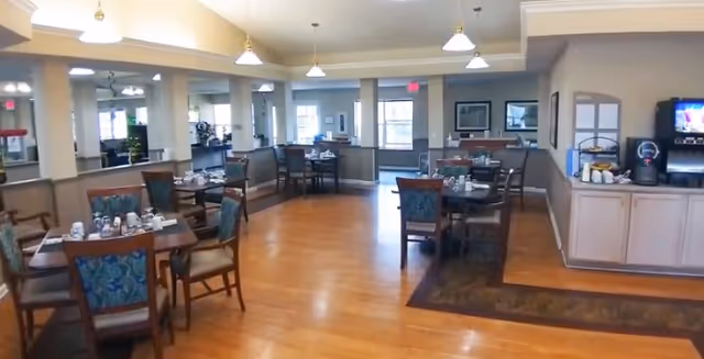 A spacious dining room with wooden floors and multiple tables set with cups and napkins. The room has several windows allowing natural light, pendant lights hanging from the ceiling, and a beverage station with a coffee machine and cups on the right side.