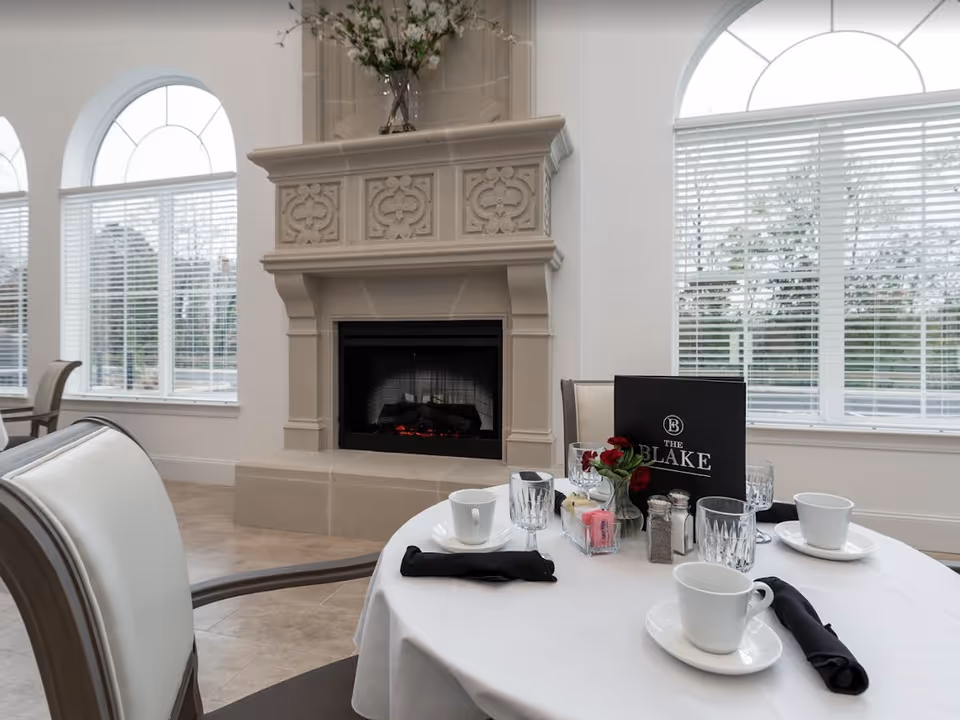 A dining area with a round table set with white cups, glasses, black napkins, and a small vase with red flowers. Behind the table is an ornate beige fireplace with a decorative mantel. Large arched windows with white blinds allow natural light into the room.