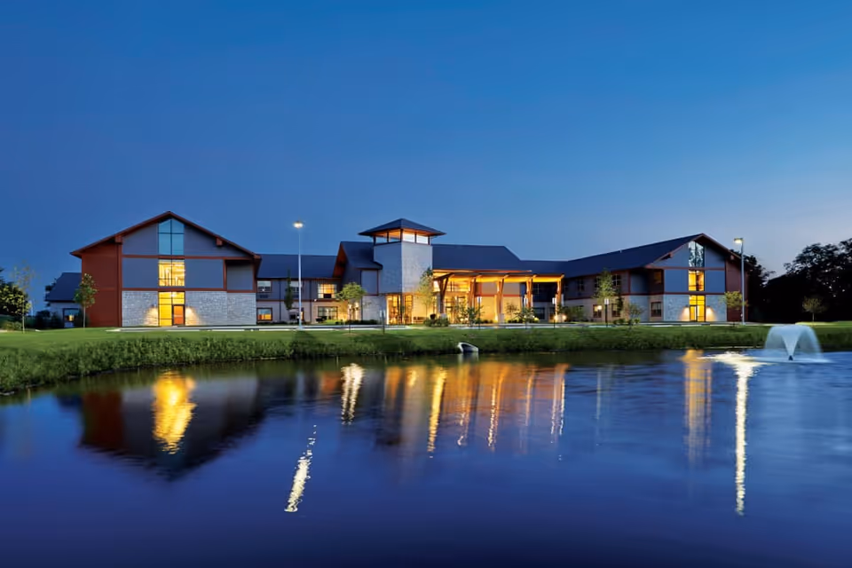 A large, modern senior living facility building illuminated at dusk with warm lights reflecting on a calm pond in the foreground, surrounded by well-maintained grass and a small fountain in the water.