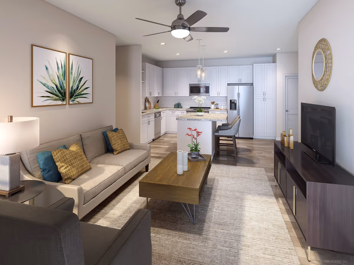 Modern open-concept living room and kitchen area in an apartment. The living room features a beige sofa with blue and gold pillows, a dark armchair, a wooden coffee table with decorative items, and a TV on a dark wooden stand. The kitchen has white cabinets, stainless steel appliances, a granite countertop island with two bar stools, and pendant lighting. The walls are light-colored with framed botanical artwork and a decorative round mirror.