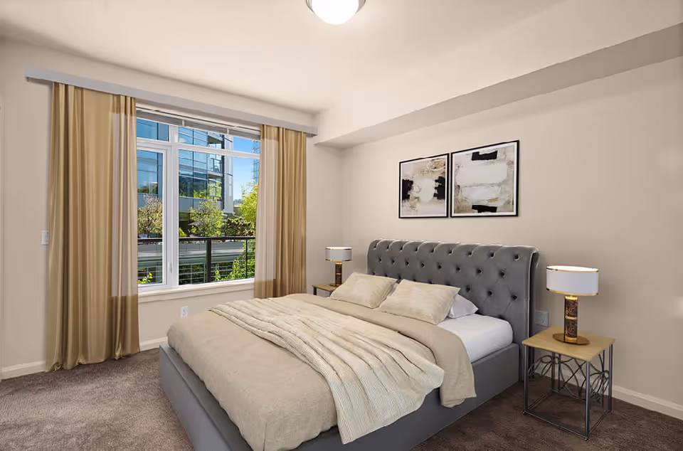 A modern bedroom with a large window covered by beige curtains, a gray upholstered bed with beige bedding and pillows, two bedside tables each with a lamp, and two abstract paintings hanging on the wall above the bed.