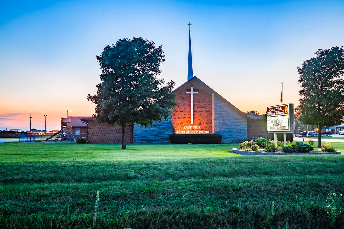Exterior view of East Side Church of the Nazarene at sunset with a large cross on the building's front wall, a tall steeple, two trees, a well-maintained lawn, and a signboard displaying church information.