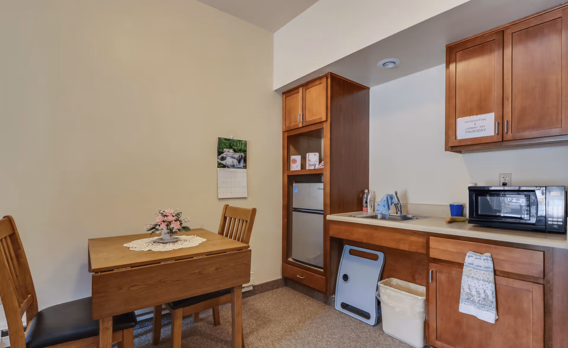 Small kitchenette area with wooden cabinets, a sink, a microwave, and a mini refrigerator. A wooden table with two chairs is placed nearby, with a small floral centerpiece on a doily. A calendar hangs on the wall above the table.