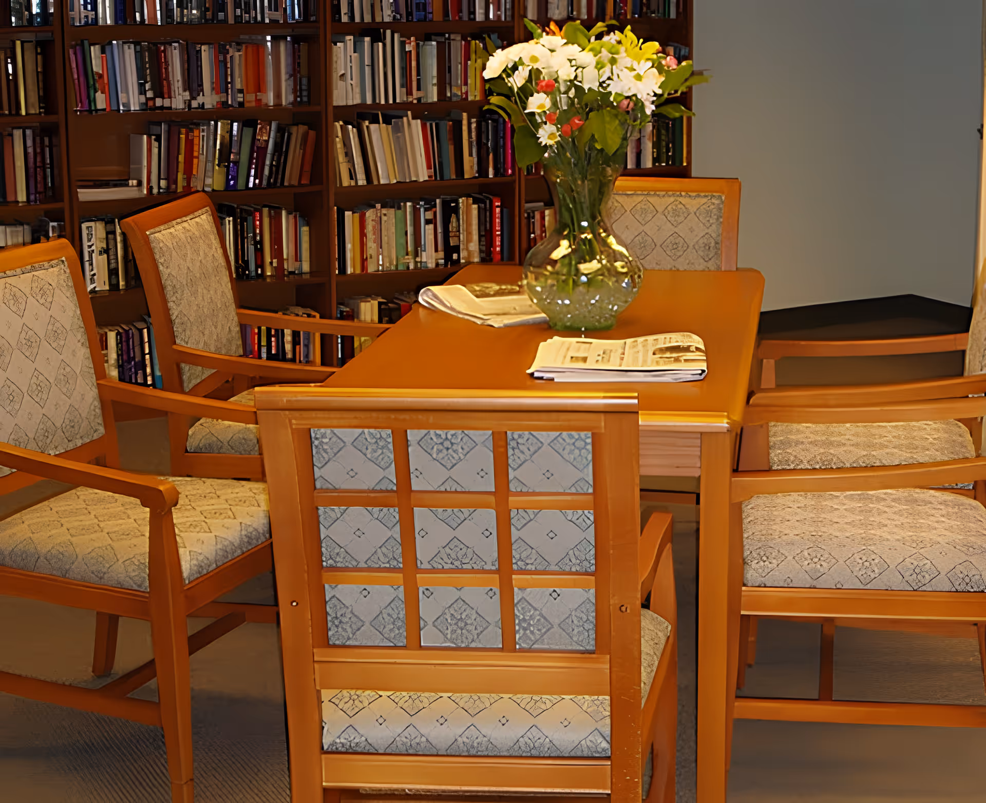 A cozy library or reading room with a wooden table surrounded by six cushioned chairs. On the table, there is a glass vase with white and red flowers and some newspapers. In the background, there are bookshelves filled with books.