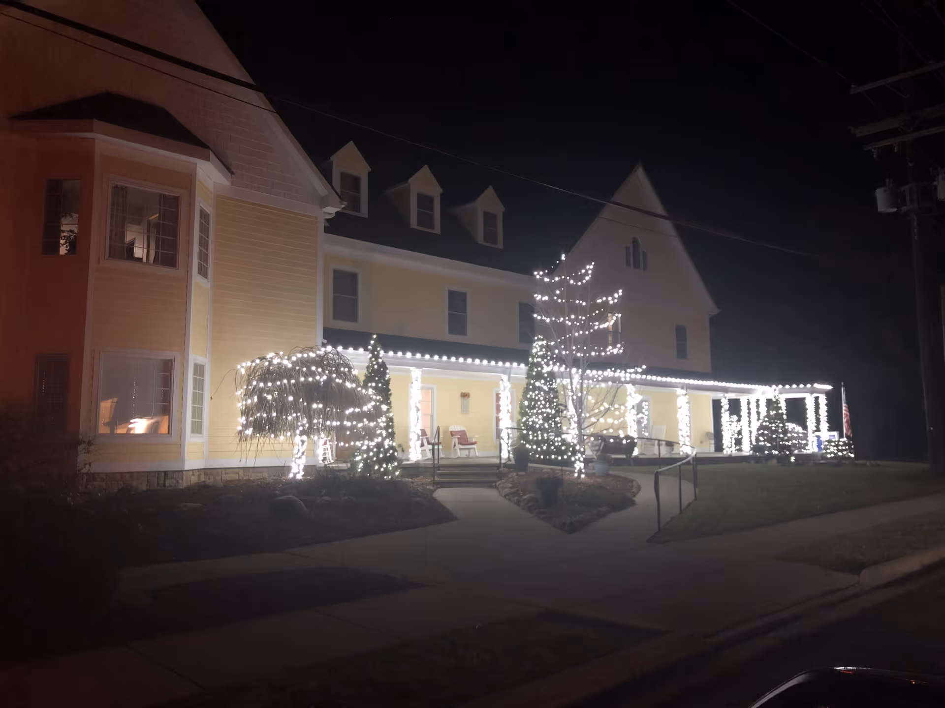 A large yellow building decorated with white string lights on trees, bushes, and along the roofline at night. The building has multiple windows and a porch with chairs, and a sidewalk leading up to the entrance.