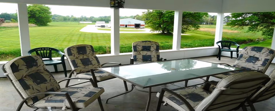 Covered porch with a glass-top patio table and cushioned chairs overlooking a grassy field and distant barn.