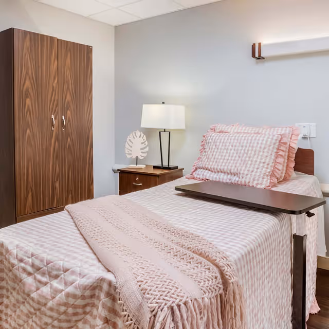 A single nursing facility bedroom with a made bed in pink gingham bedding, an overbed tray, bedside lamp and a wooden wardrobe.