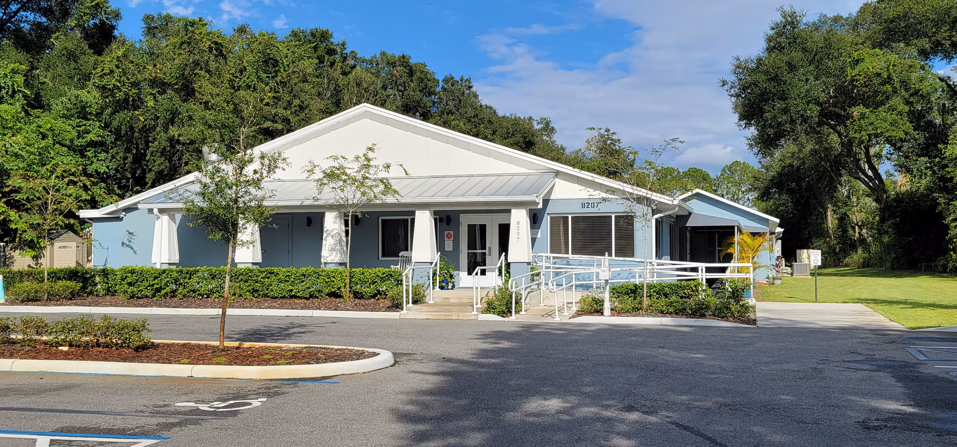 Single-story light blue building with a white roof and a wheelchair ramp at the entrance, surrounded by greenery and trees under a partly cloudy sky. There is a parking lot in front with marked spaces.