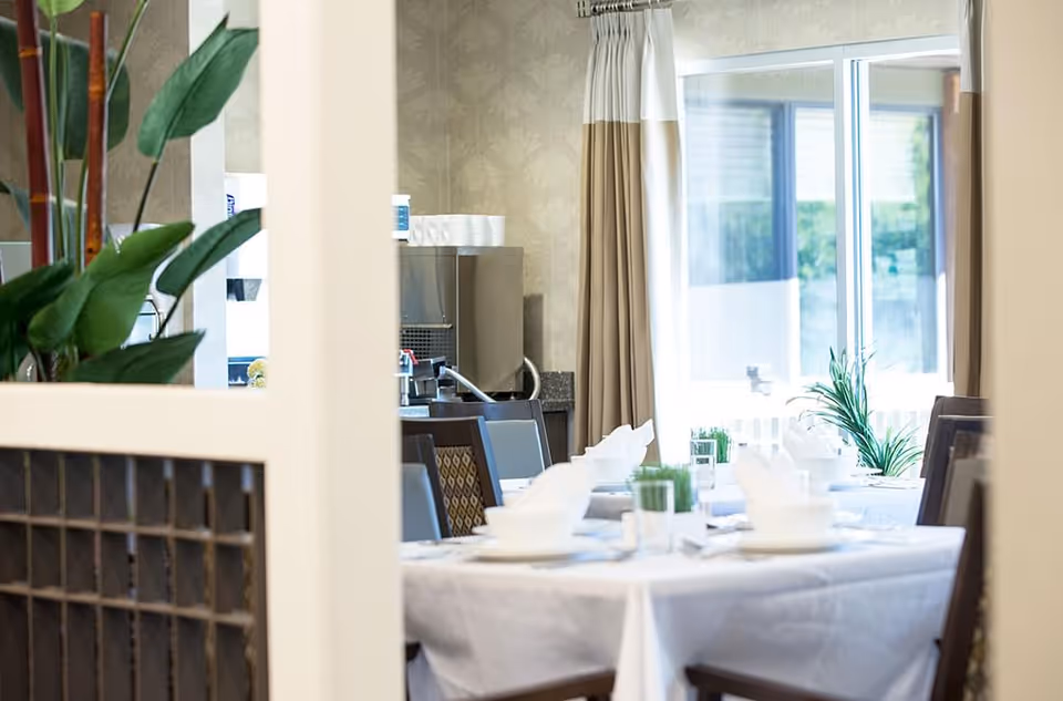 View through a partition into a dining room with a table set for a meal, including white bowls, plates, and napkins. There are chairs around the table, potted plants as centerpieces, and large windows with beige curtains letting in natural light.