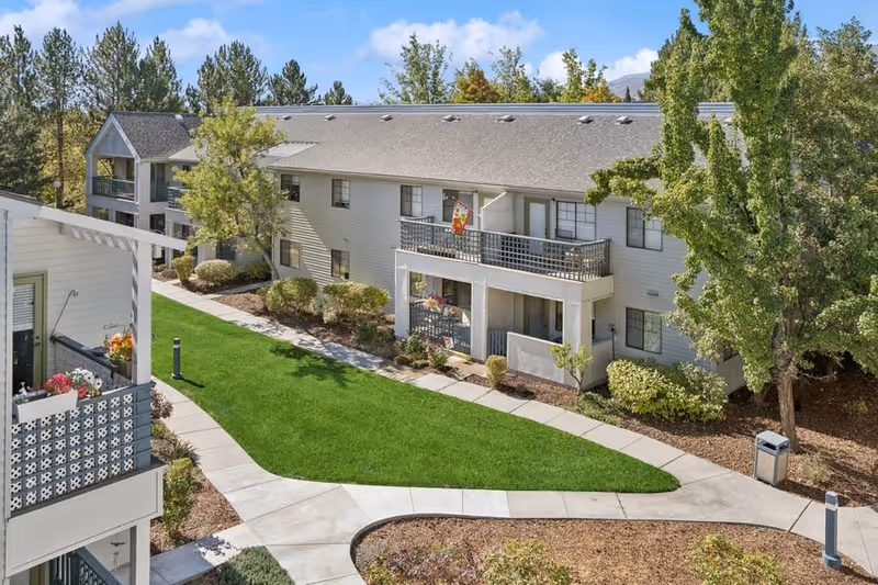 Exterior view of a two-story residential building with balconies and patios, surrounded by green lawns, trees, and landscaped pathways under a blue sky.