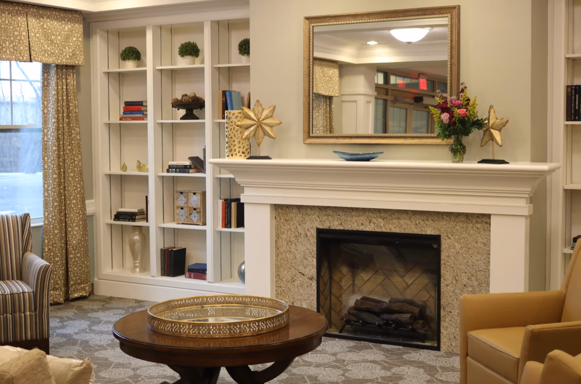 A cozy living room area in a senior living facility featuring a fireplace with a granite surround and white mantel. On the mantel are decorative items including a gold star sculpture, a vase with colorful flowers, and a blue dish. Above the fireplace is a large gold-framed mirror reflecting part of the room. To the left, there is a built-in white bookshelf with various books and decorative objects, and a window with patterned curtains. In front of the fireplace is a round wooden coffee table with a decorative gold tray. There are upholstered chairs in striped and solid patterns around the room.