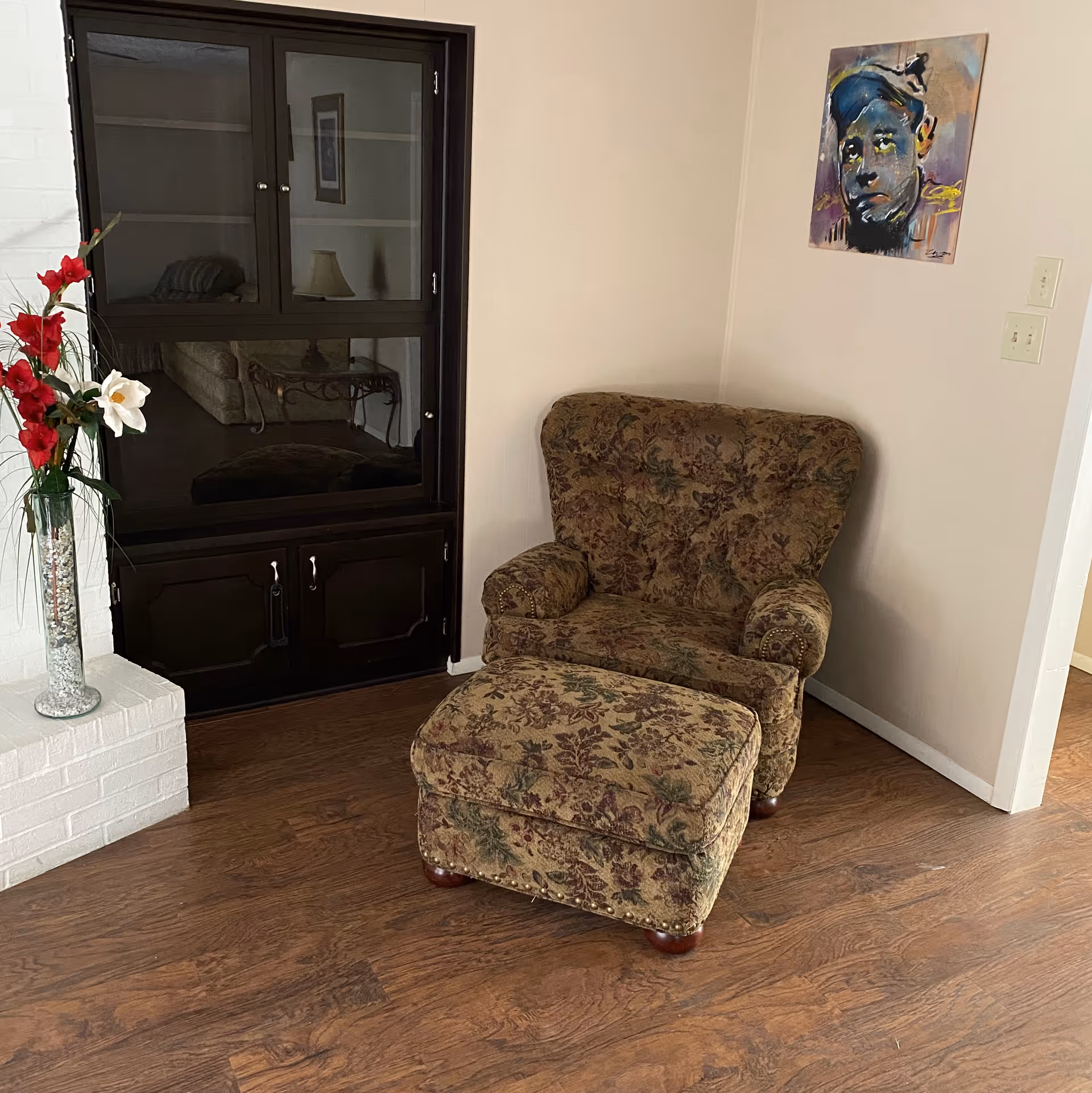 A cozy corner of a living room featuring a floral-patterned armchair with a matching ottoman on a wooden floor. To the left, there is a white brick structure with a tall glass vase holding red and white flowers. Behind the chair is a dark wooden cabinet with glass doors reflecting another room with a lamp and sofa. A colorful portrait painting hangs on the wall above the chair.