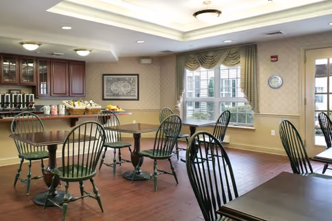 A dining room with several wooden tables and green wooden chairs. The room has large windows with patterned curtains allowing natural light to enter. There is a counter area with coffee dispensers, cups, and snacks. The walls are decorated with wallpaper and a framed picture. The floor is wooden, and ceiling lights illuminate the space.