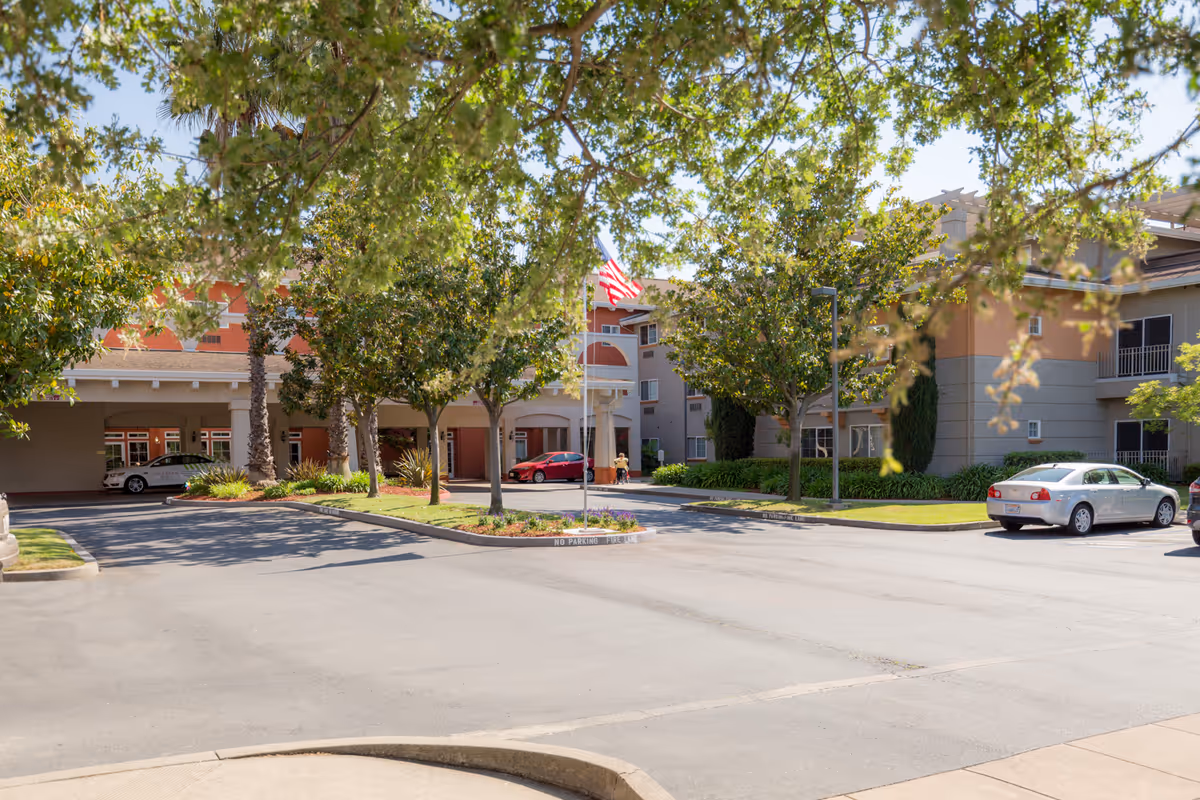 Exterior view of Cogir of Stock Ranch facility showing a driveway with parked cars, trees, and a building entrance with an American flag on a flagpole.