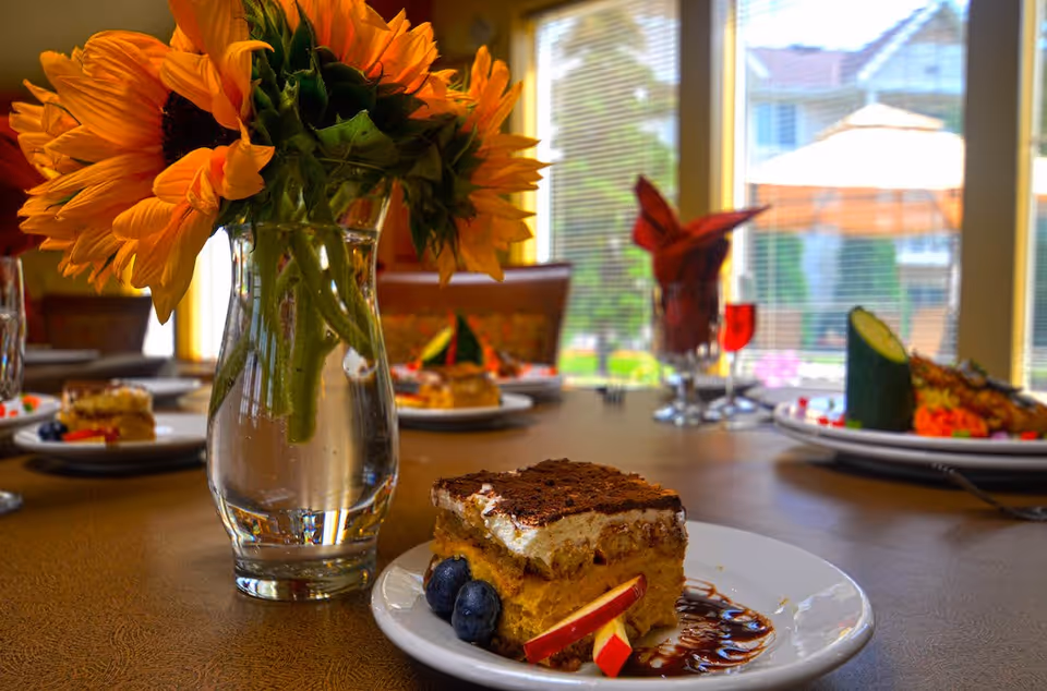 A close-up view of a dessert plate with a slice of cake garnished with blueberries and apple slices, placed on a table next to a vase of sunflowers. In the background, there are other plates with food and a window showing an outdoor scene with umbrellas and greenery.