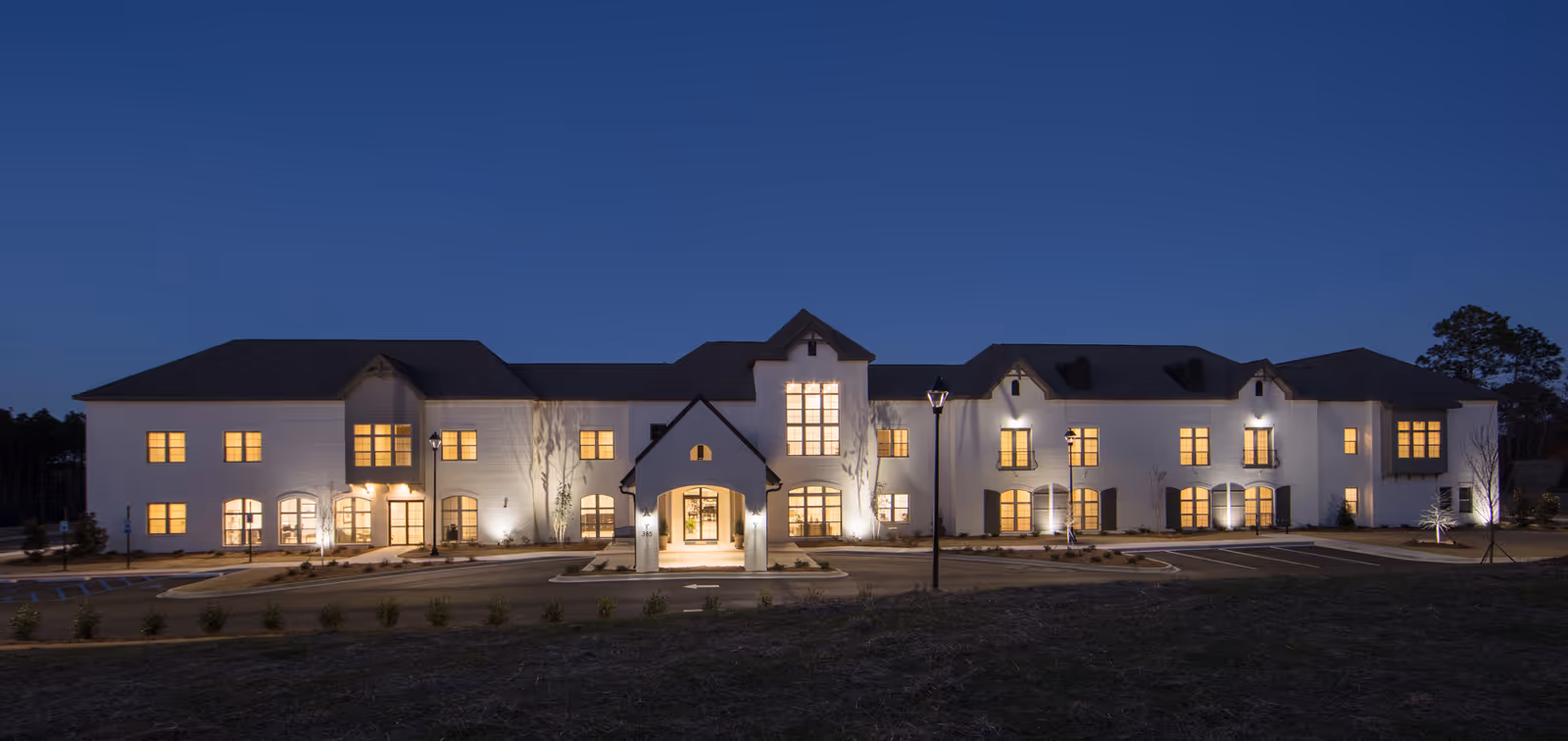 Exterior view of The Blake at Edgewater senior living facility at dusk, showing a large two-story building with multiple lit windows and a central entrance under a peaked roof. The building is white with dark roofing, surrounded by a paved driveway and some landscaping.