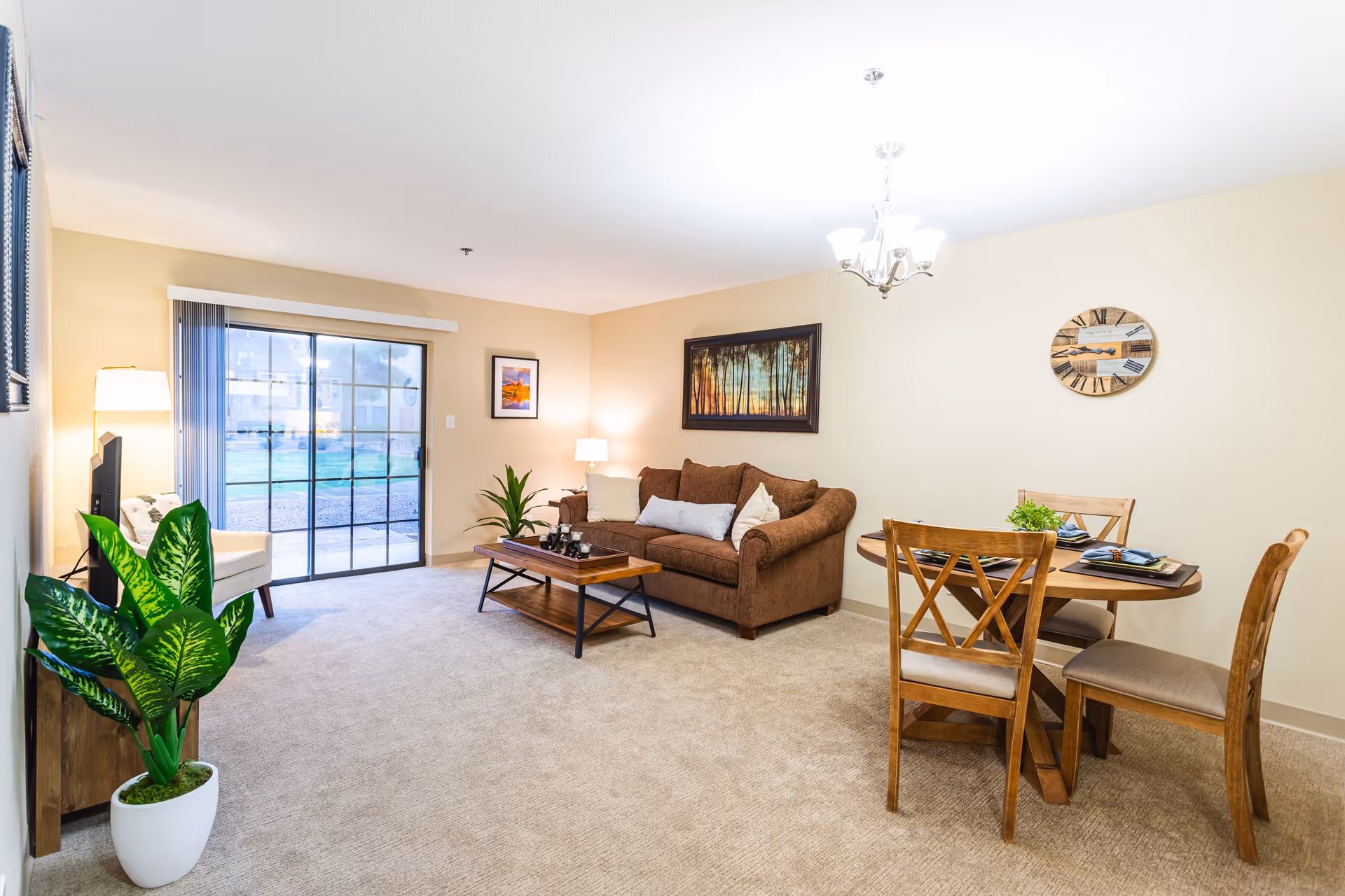A bright and cozy living room with beige walls and carpet. The room features a brown sofa with cushions, a wooden coffee table, a round wooden dining table with four chairs, a potted plant, a floor lamp, and framed artwork on the walls. Large sliding glass doors lead to an outdoor area.