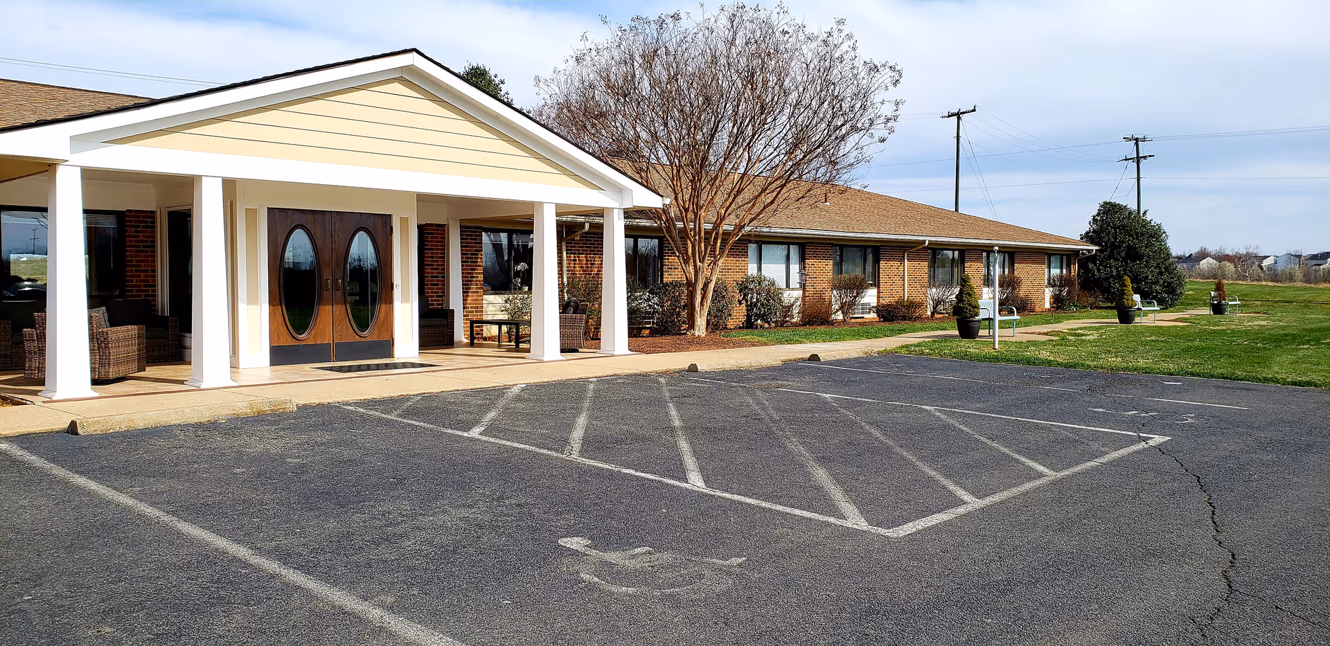 Exterior view of a single-story brick building with a covered entrance supported by white columns. There are double wooden doors with oval glass panels at the entrance, outdoor seating with wicker chairs, a parking lot with a handicapped parking space, and some trees and shrubs around the building under a partly cloudy sky.