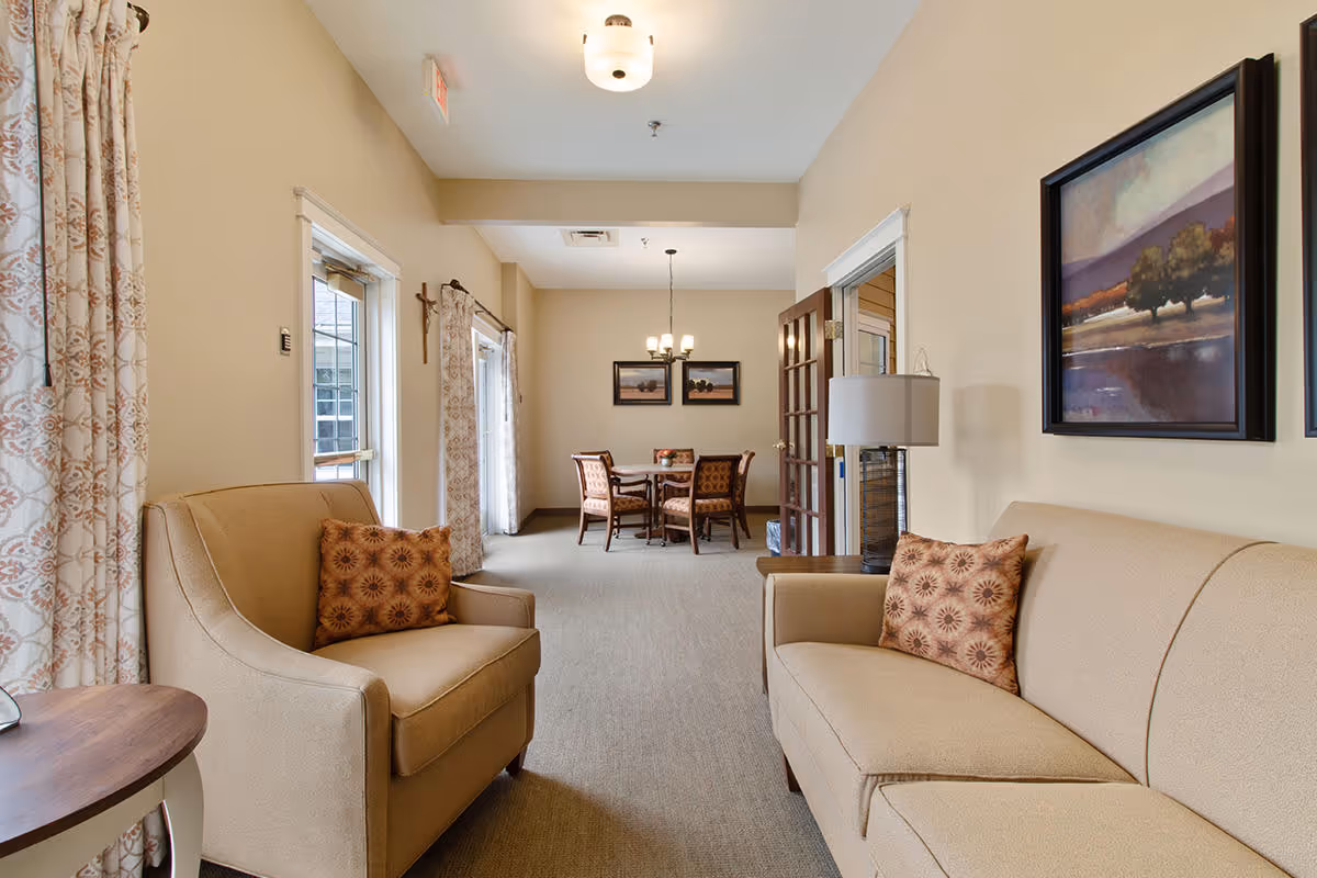 A cozy senior living common area with beige walls and carpet. The foreground features a beige armchair and sofa, each with a patterned throw pillow. A wooden side table with a lamp is next to the sofa. In the background, there is a dining area with a round table and four chairs under a chandelier. Two framed landscape paintings hang on the wall above the dining table. Light filters in through windows with patterned curtains on the left side.