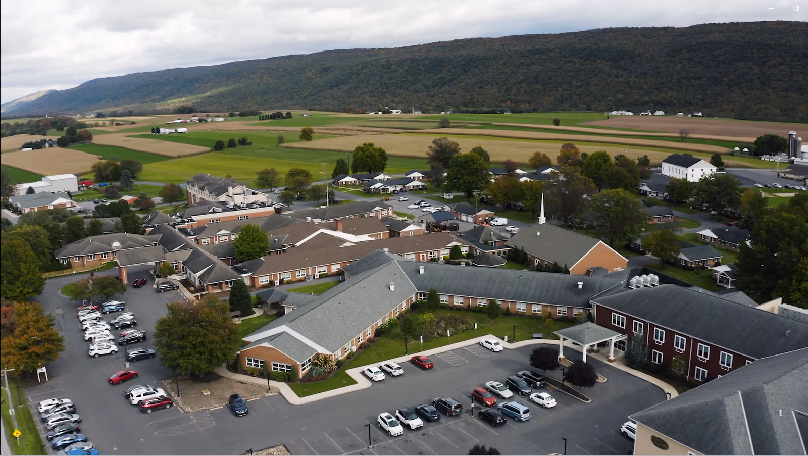 Aerial view of Valley View Retirement Community showing multiple connected buildings with parking lots, surrounded by green fields and hills in the background under a cloudy sky.